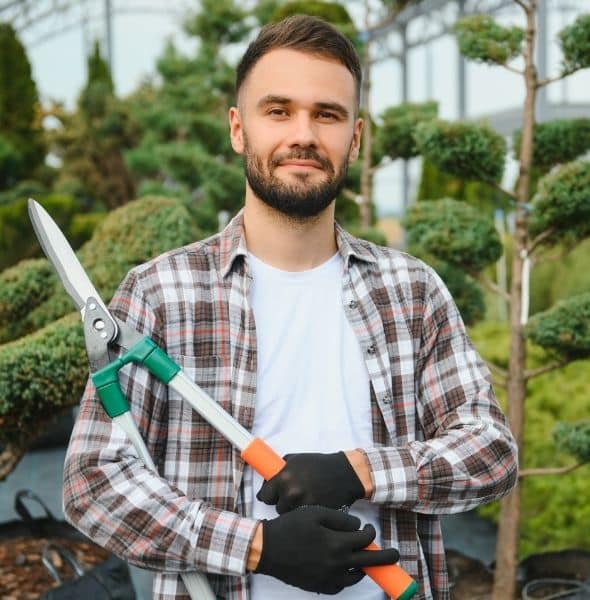 Paysagiste tenant un taille-haie manuel devant des arbustes soigneusement entretenus dans un jardin paysager.